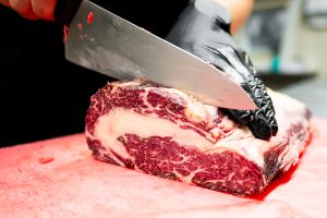 UV decontamination solutions for the meat processing industry. Gloved hand slicing raw steak on a red cutting board in a butcher or meat processing facility, illustrating UV decontamination solutions for knives, conveyor belts and air.